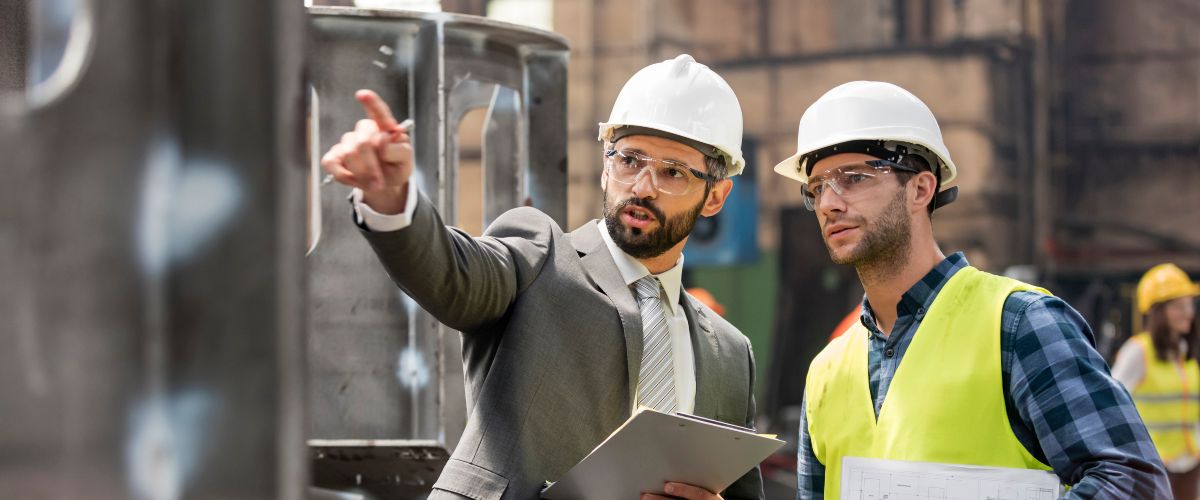 Symbolbild: Zwei Männer mit Bauarbeiterhelmen stehen vor Industrieanlage