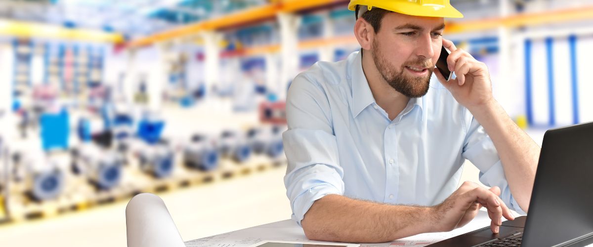 Ein Ingenieur sitzt in einer Fabrikhalle vor seinem Laptop und telefoniert mit dem Handy (Symbolbild).